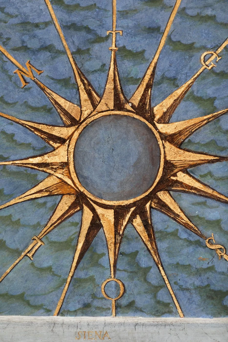 A painted artistic depiction of a compass rose, with a central circle and radiating cardinal points marked N, E, S, W, on a cloudy blue background. The word 'SIENA' is written at the bottom.