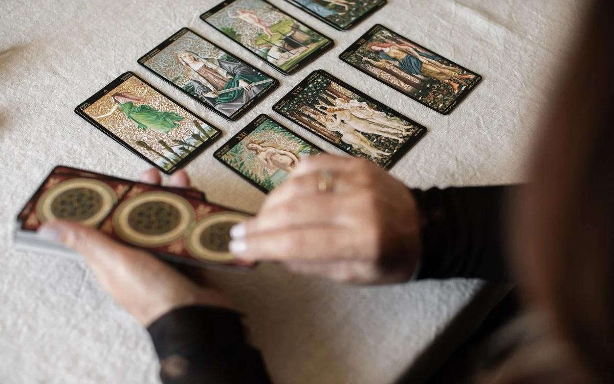 A person with long hair and a black sleeve is holding a deck of tarot cards, while several other tarot cards are spread out on a light-colored surface.