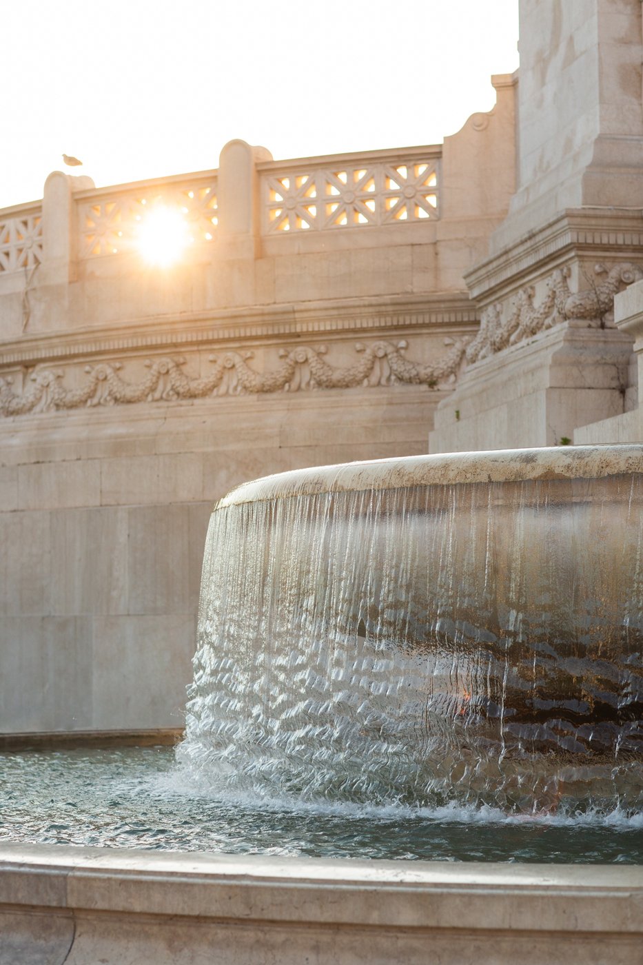 A decorative fountain with water cascading over the edge, situated in front of a stone building with ornate carvings and a balustrade, illuminated by the setting sun.