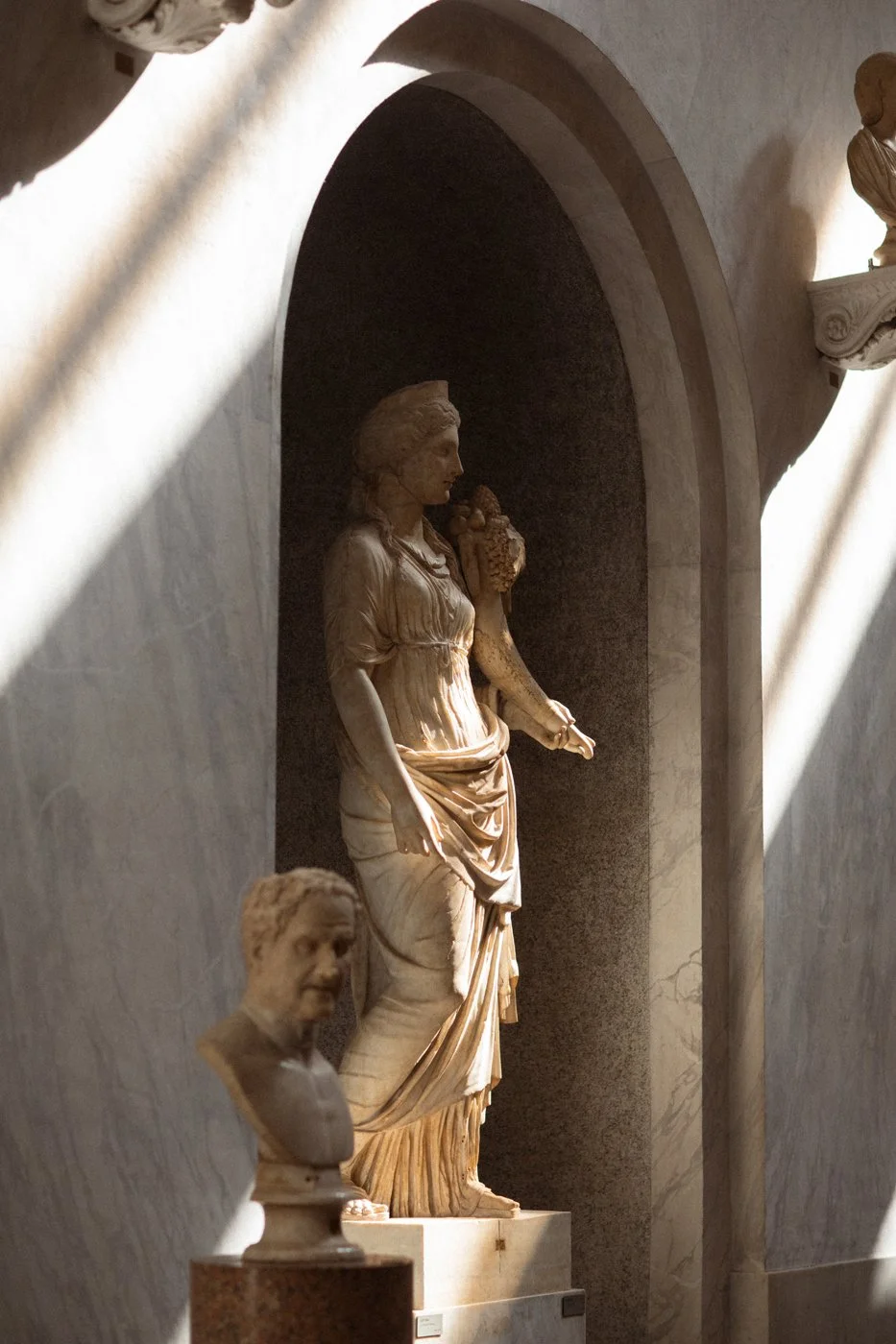 A classical marble sculpture of a woman standing in a niche with sunlight streaming in. A bust sculpture is in the foreground.