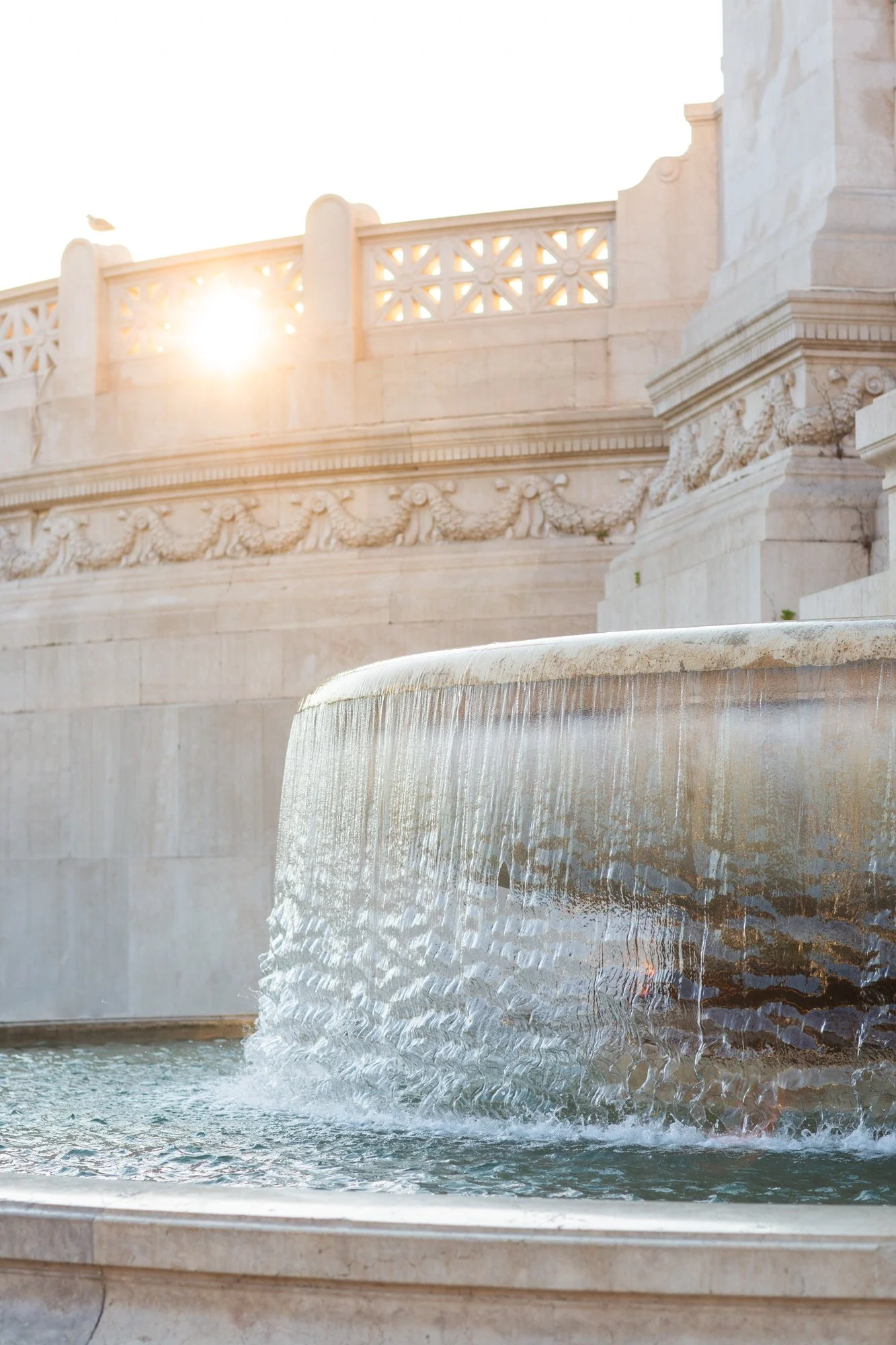 A white marble fountain with water cascading over a wide, curved basin, located in front of a historic building with intricate carvings and decorative motifs, illuminated by sunlight.