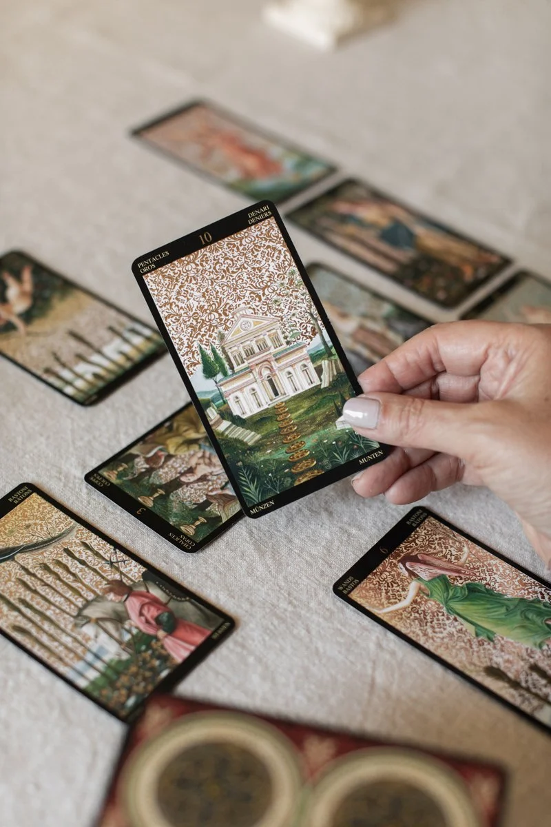 A hand holding a tarot card depicting a large mansion with a golden staircase, surrounded by trees and set against a patterned background. Several other tarot cards are laid out on a light-colored surface.