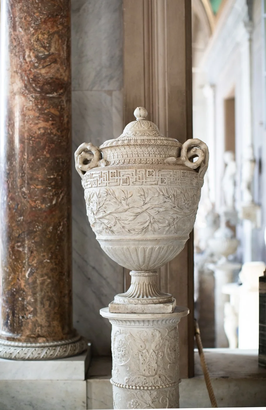 Ancient Greek or Roman marble urn with intricate carvings, handles, and a lid, displayed on a decorative column.