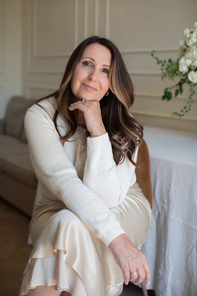 A woman with long brown hair sitting on a chair in a room with cream-colored walls and a beige sofa. She is wearing a cream-colored sweater and a matching skirt, with a ring on her left hand and earrings. She is resting her chin on her left hand and smiling softly at the camera. There is a bouquet of white flowers on a table behind her.