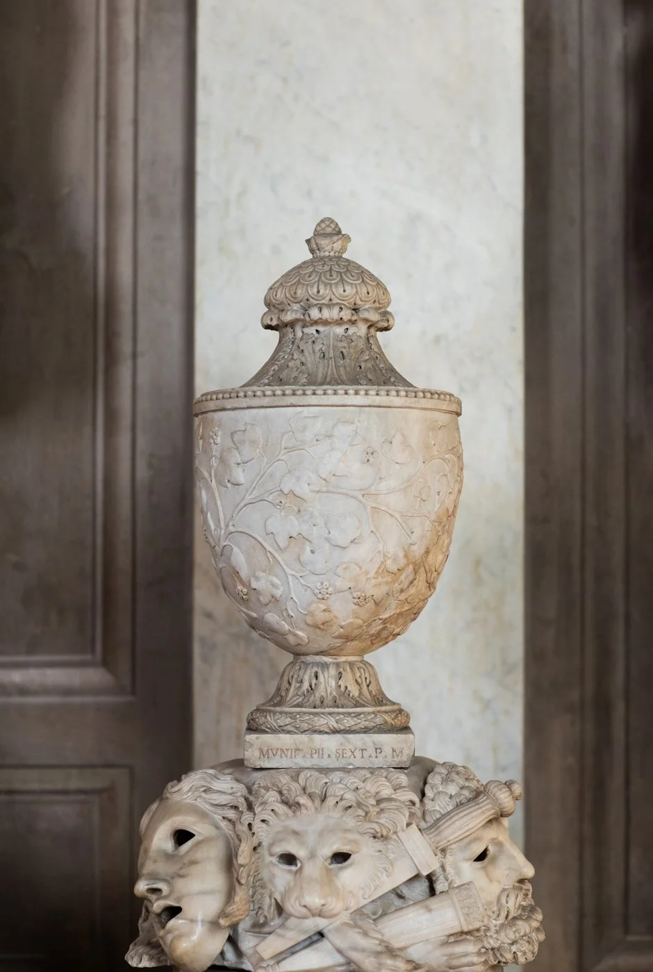 Marble sculpture featuring a detailed urn with floral carvings, placed on a pedestal surrounded by carved heads of human faces with distinctive expressions.