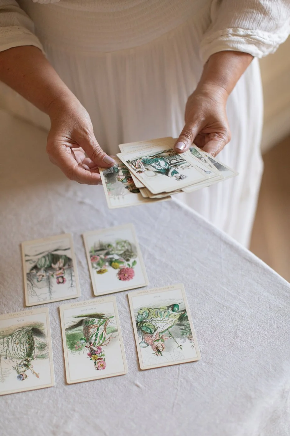 Person dressed in white is holding a spread of tarot or oracle cards, with more cards laid out on a table covered with a white cloth.