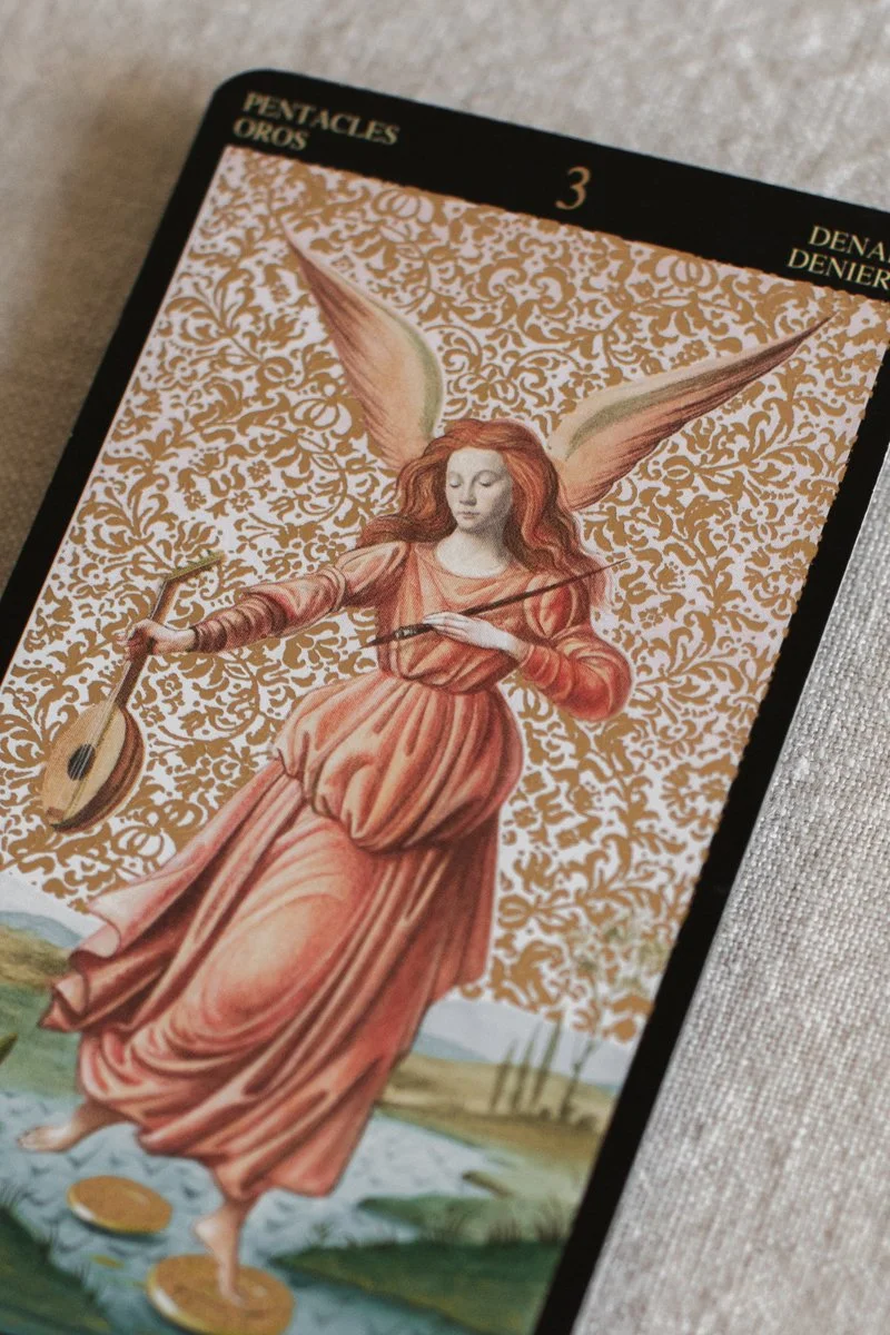 Tarot card titled 'Pentacles Oros' with the number 3. It depicts a woman with long, flowing red hair, wearing a pink dress, holding a bow in her left hand and an arrow in her right hand. She has large angelic wings and stands on a body of water, with a golden ornate background.