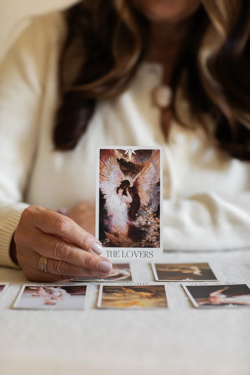 A woman holding up a tarot card titled "The Lovers" with other tarot cards spread on the table in front of her.