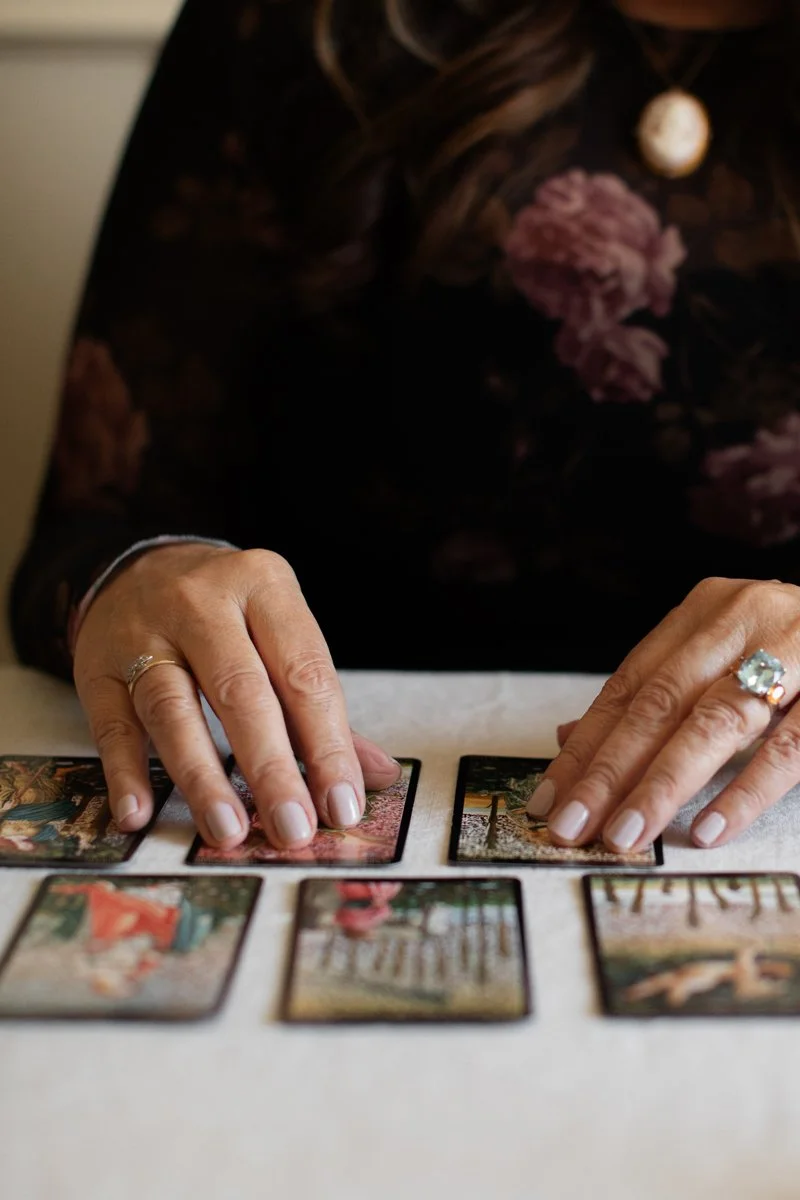 Person laying out tarot or playing cards on a table, wearing rings, with long curly hair, in a dark floral top.