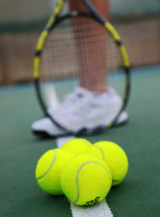 Close-up of tennis balls on a tennis court with a tennis racket and a player's foot wearing white sneakers in the background.