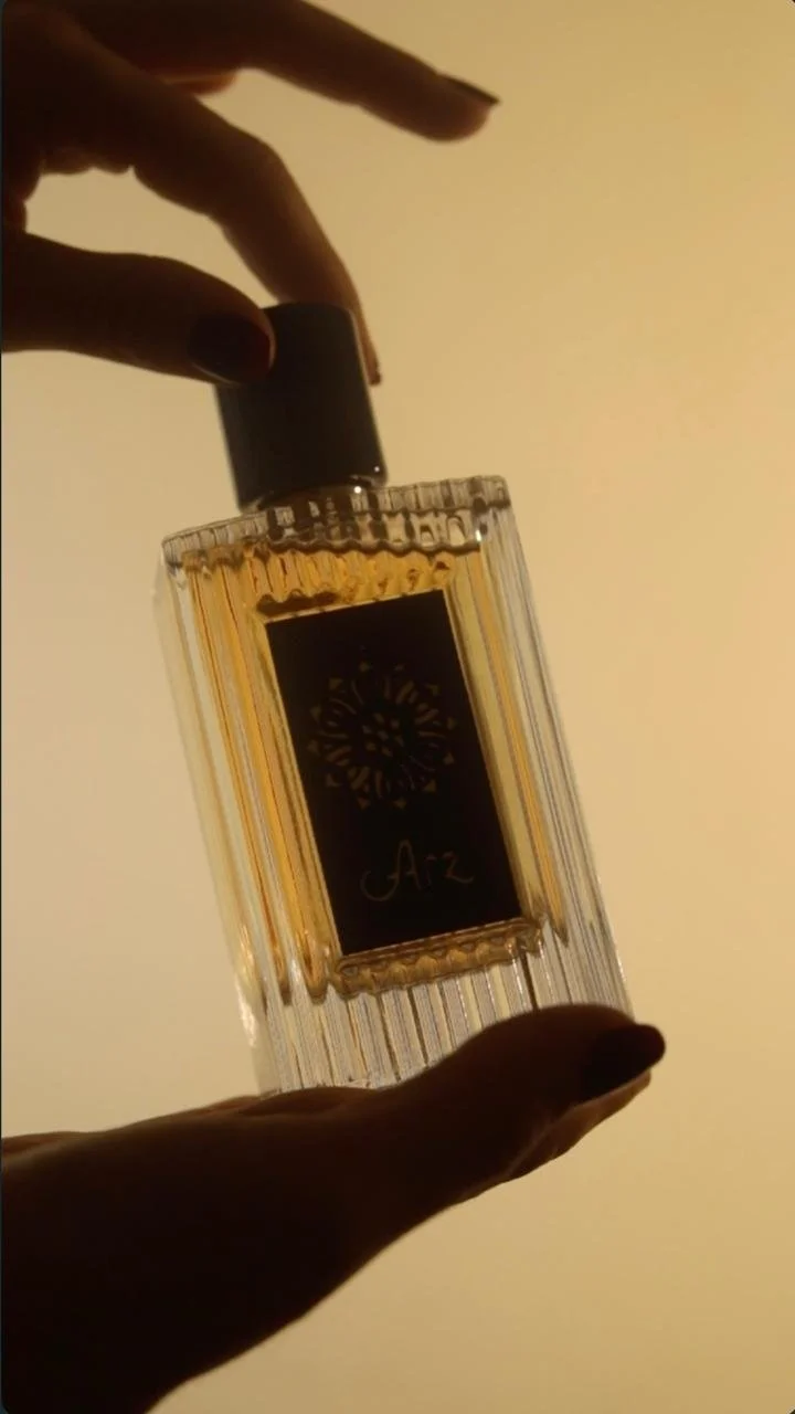 A person's hand holds a bottle of Azz perfume with a gold and black label. The bottle is rectangular with vertical ridges, and the photograph is taken against a plain background with warm lighting.
