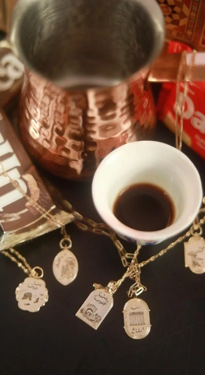 A metallic pitcher, a cup of black coffee, and gold jewelry with Arabic writing and designs on a dark surface.