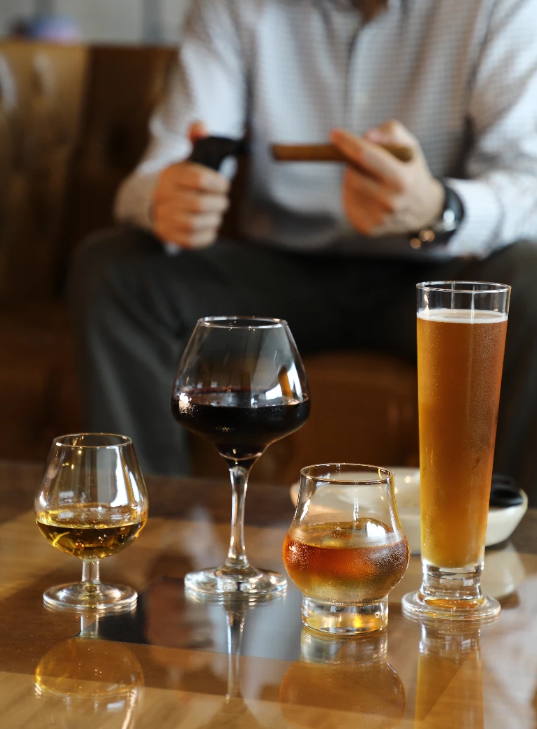 Four glasses of different types of alcohol on a table with a person sitting in the background holding a cigar.