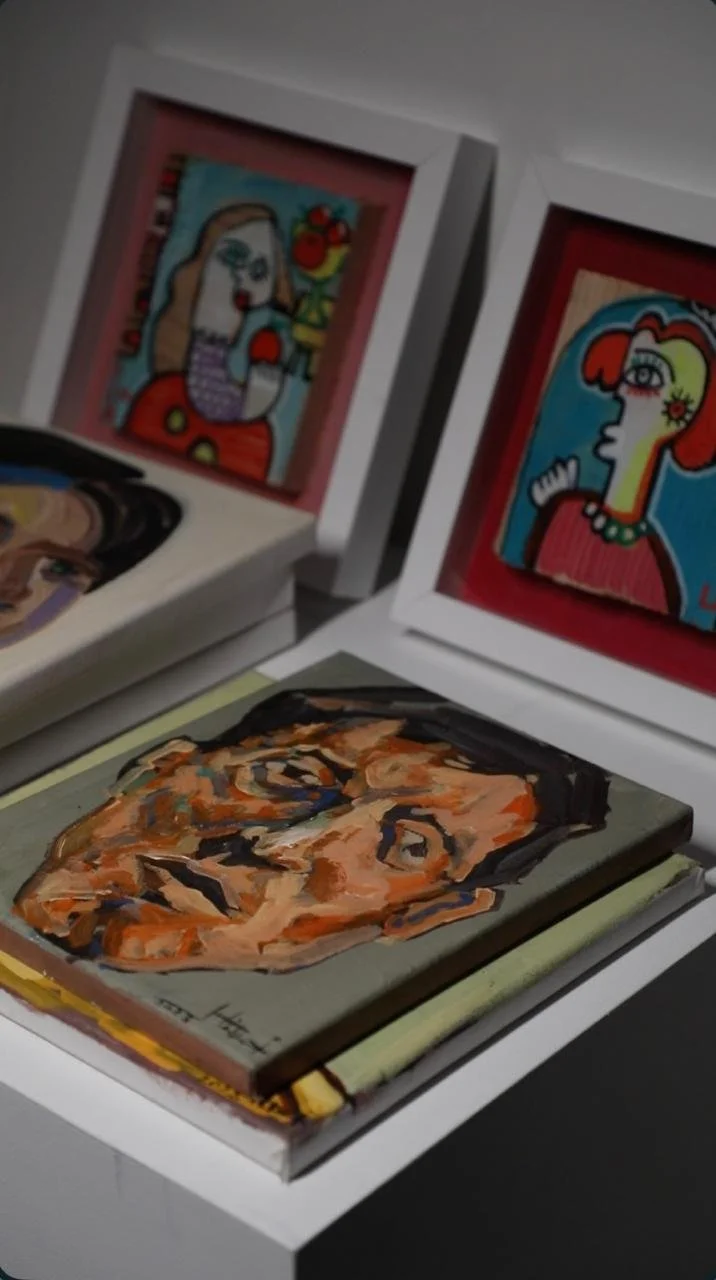 A collection of art books and framed colorful illustrations, including a painting of a woman with flowers in her hair and a bird, and another portrait with exaggerated facial features.