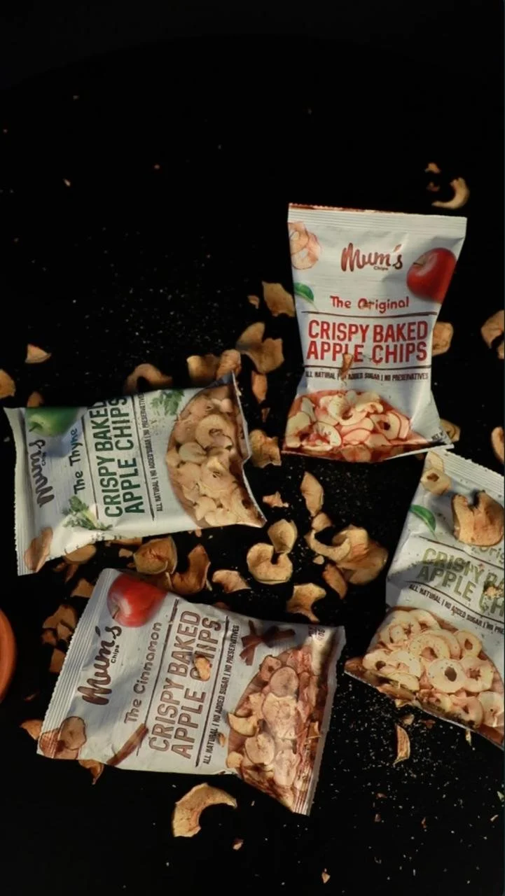 Various bags of Mum's crispy baked apple chips spilled on a black surface with scattered apple chips around.