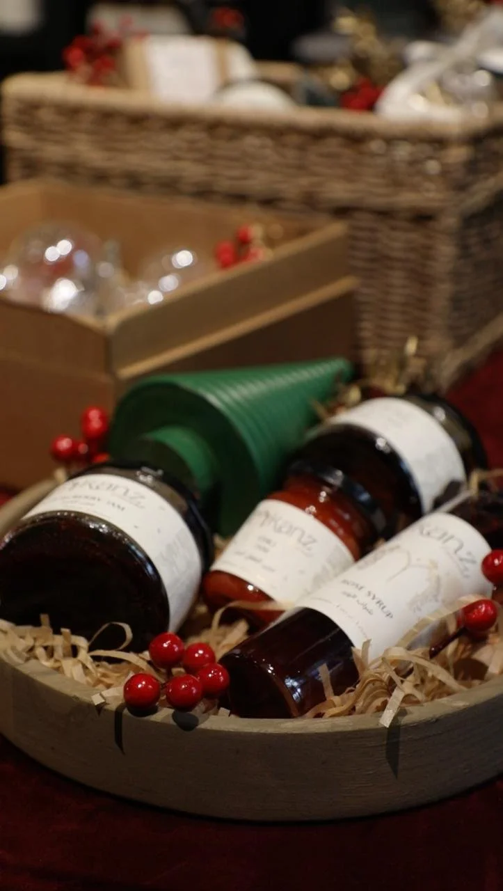 Holiday gift basket containing jars of jam, a green Christmas tree-shaped item, red berries, and a basket filled with wrapped presents in the background.