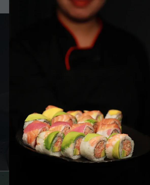 A person holding a black plate with assorted sushi rolls, topped with slices of salmon, tuna, yellowtail, and avocado, against a black background.