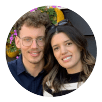 Due persone sorridenti che posano insieme, un uomo con occhiali e una donna con capelli scuri chiari, sotto un arco decorato con fiori e colori vivaci.