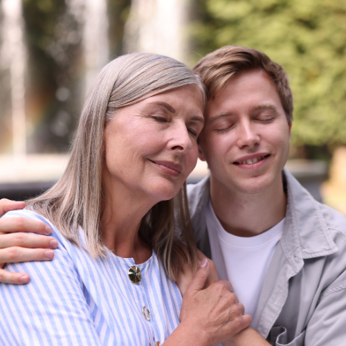 Una donna anziana e un giovane uomo si abbracciano con gli occhi chiusi in un parco, mostrando emozioni di affetto e serenità.