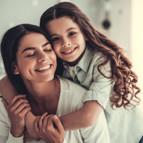 Una donna e una bambina sorridenti abbracciate, in un ambiente domestico luminoso.