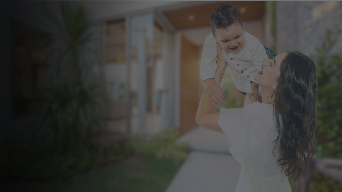Una donna con capelli lunghi e scuri solleva un bambino in aria, entrambi sorridono. Sono all'aperto, in giardino, con piante e una struttura in legno sullo sfondo.