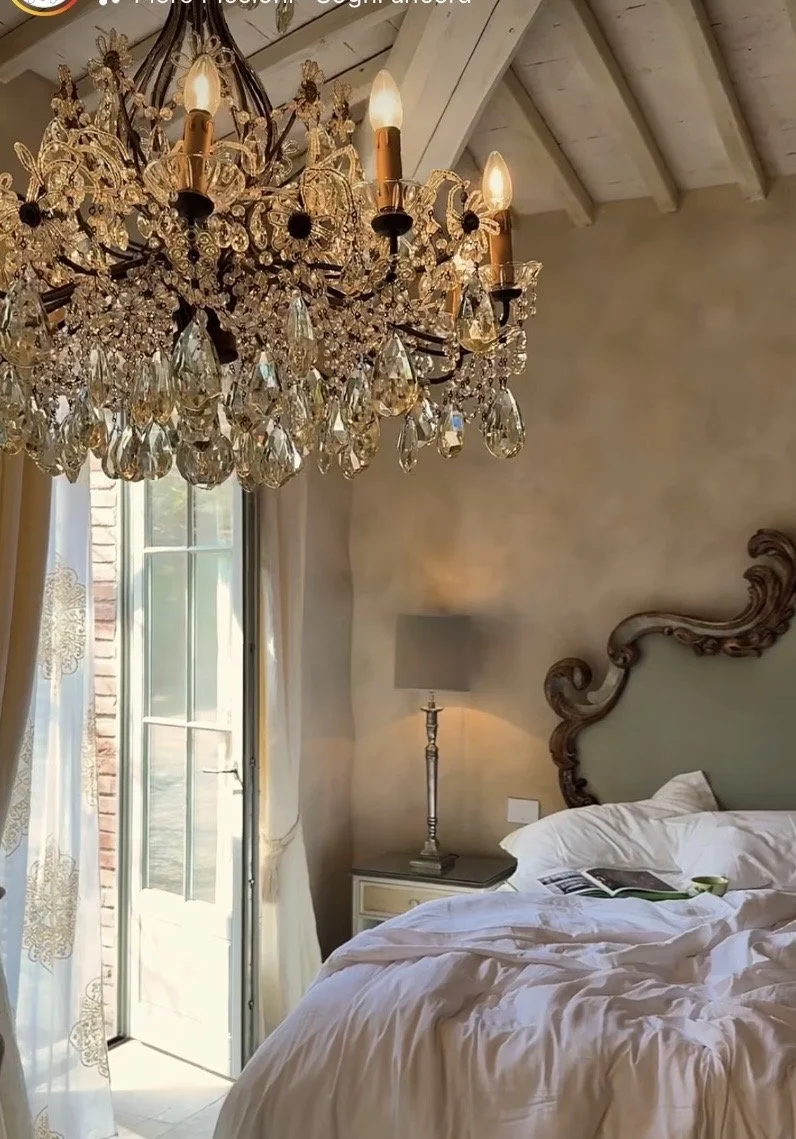 Luxurious bedroom with an ornate crystal chandelier, elegant carved headboard, white linens, and soft natural light filtering through sheer curtains.