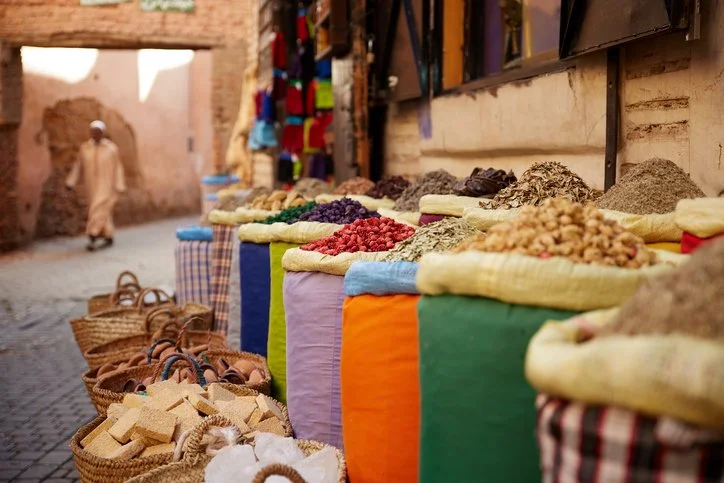 Colorful market scene with baskets and sacks of spices and nuts in the foreground. A person in traditional attire walks near rustic walls in the background.