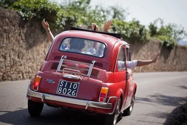 A red vintage car drives down a sunny road with the top down. Two people inside raise their arms joyfully.