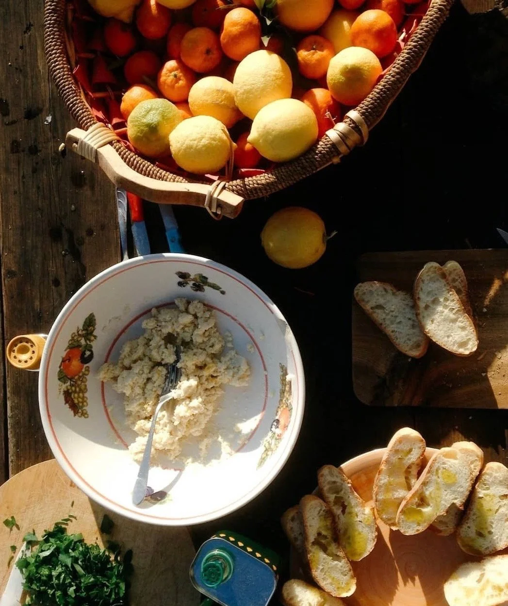 A rustic kitchen table displays bread, a bowl of oats, and a wicker basket filled with fruit.