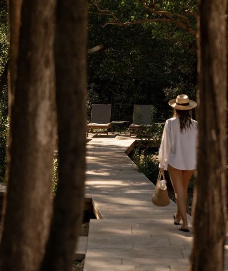 A woman in a straw hat carrying a woven bag, walking toward two empty loungers on a sunlit, tree-lined path.