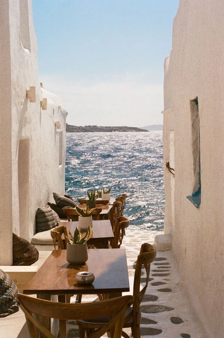 Narrow outdoor cafe with wooden tables and chairs lined along white walls, leading to a view of a sparkling blue sea under a clear sky.