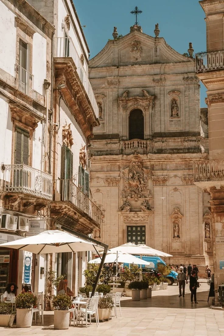 A picturesque European street showcases ornate classical architecture with a grand church facade. People stroll by outdoor cafes under white umbrellas.