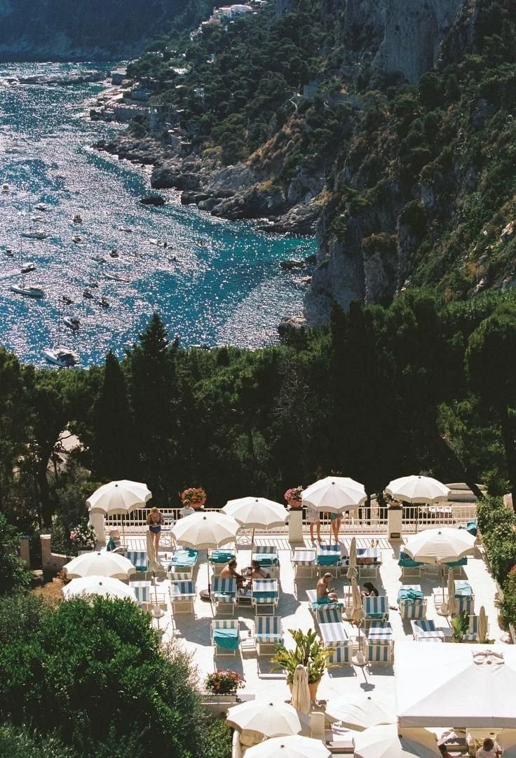 Aerial view of a coastal resort with blue ocean waters at the top, white umbrellas, and sunbathers relaxing on loungers below.