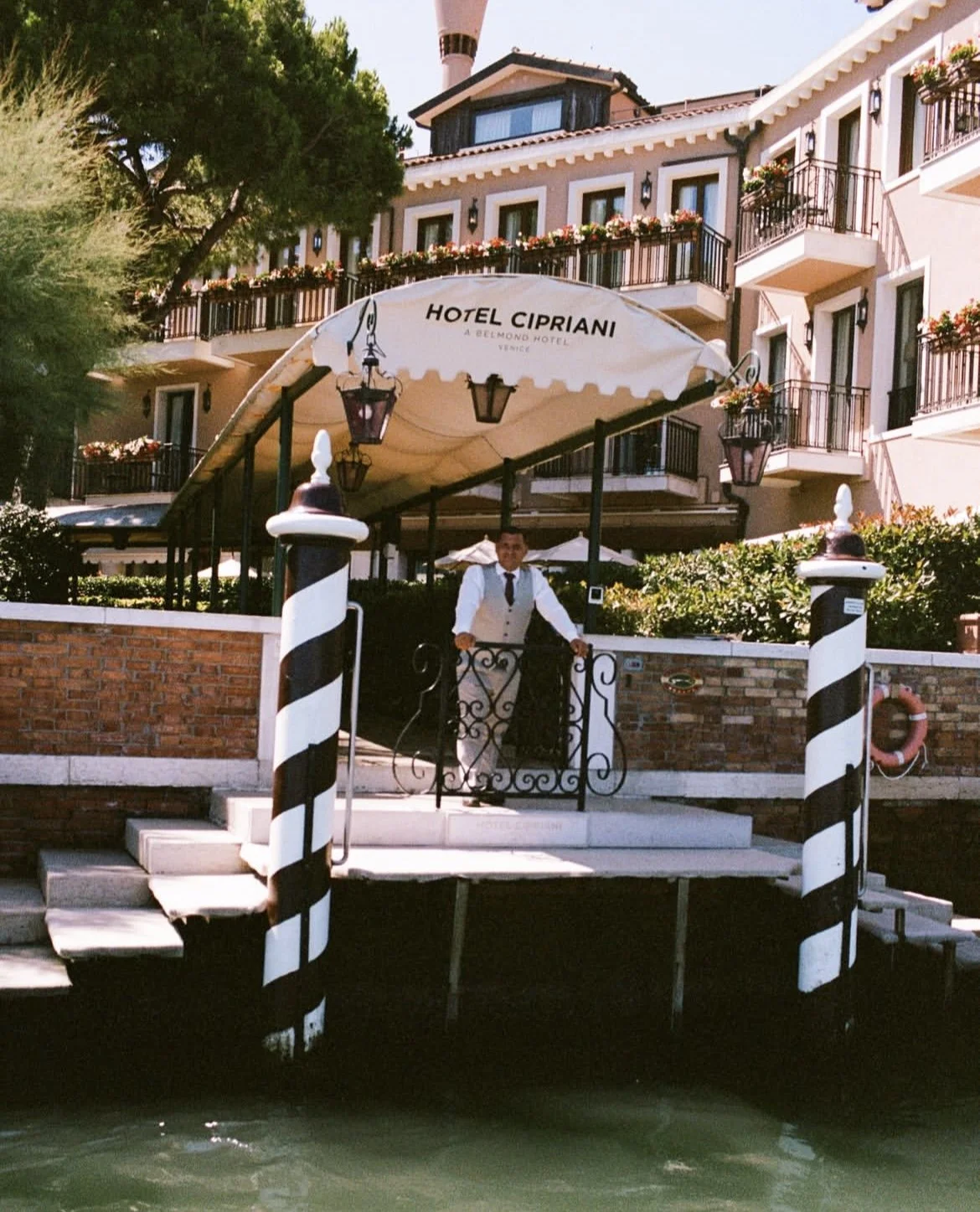 Entrance to Hotel Cipriani with a welcoming staff member. Two striped poles frame a decorative gate.