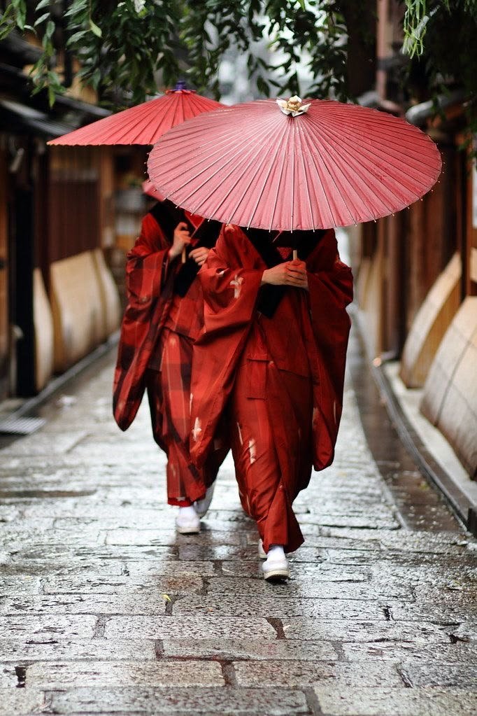 People in a red clothing walk down a narrow, wet street, with large red umbrellas.