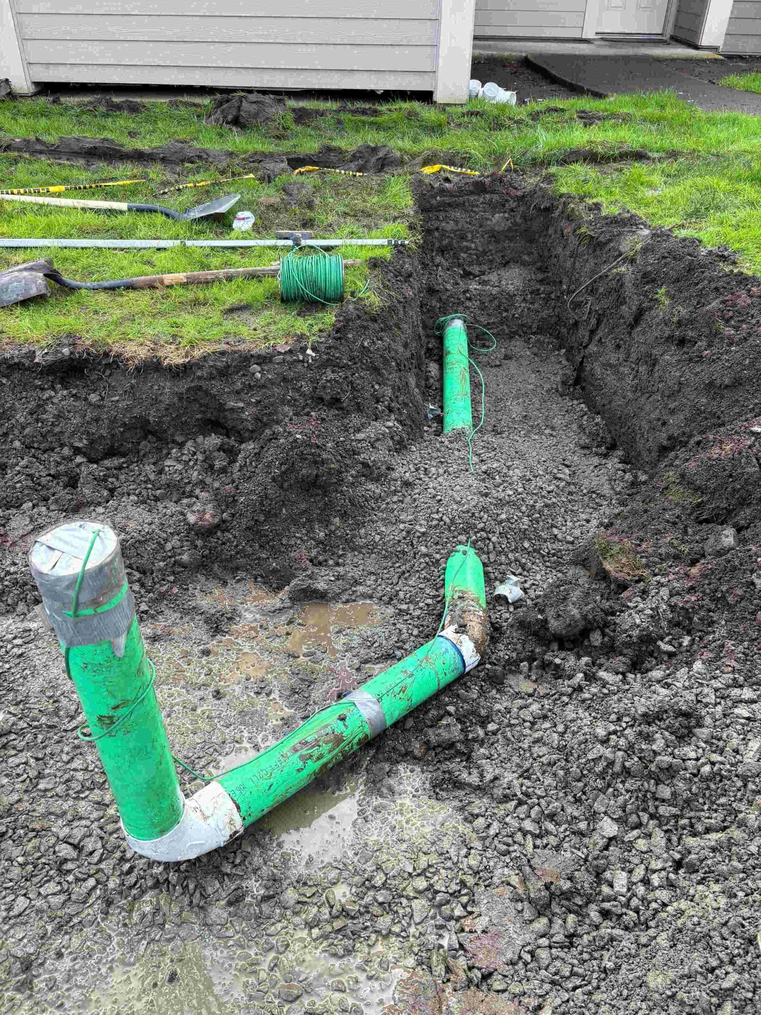 Underground plumbing installation with green pipes and construction tools in a backyard excavation site.