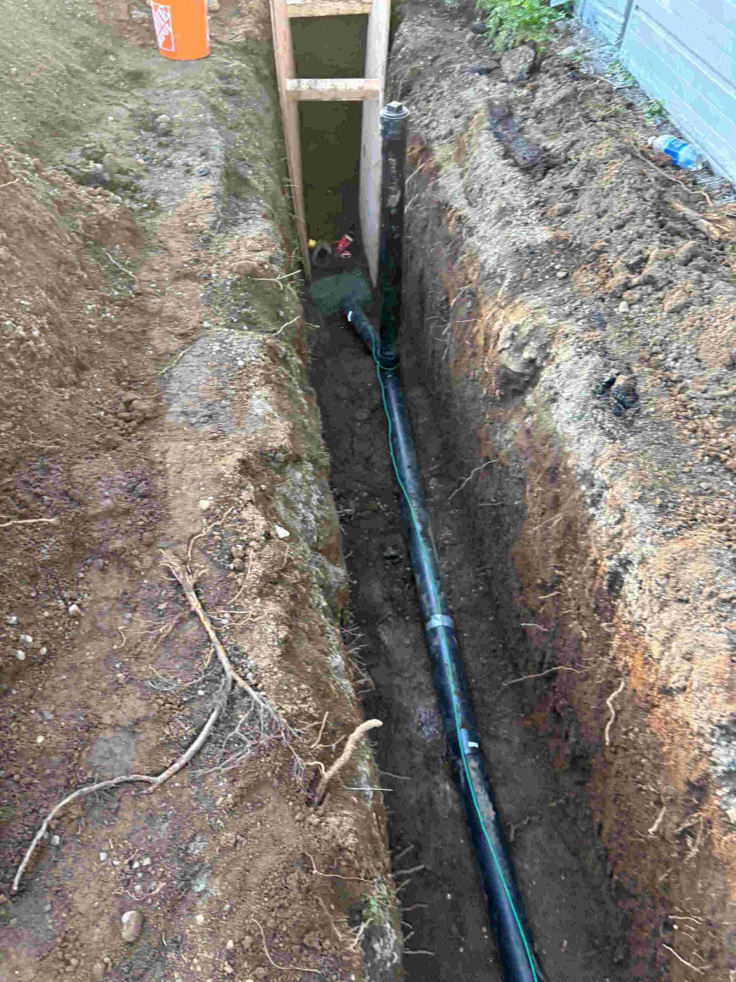 A narrow trench with an underground pipe and wiring, surrounded by dirt and soil, near a building with blue siding.