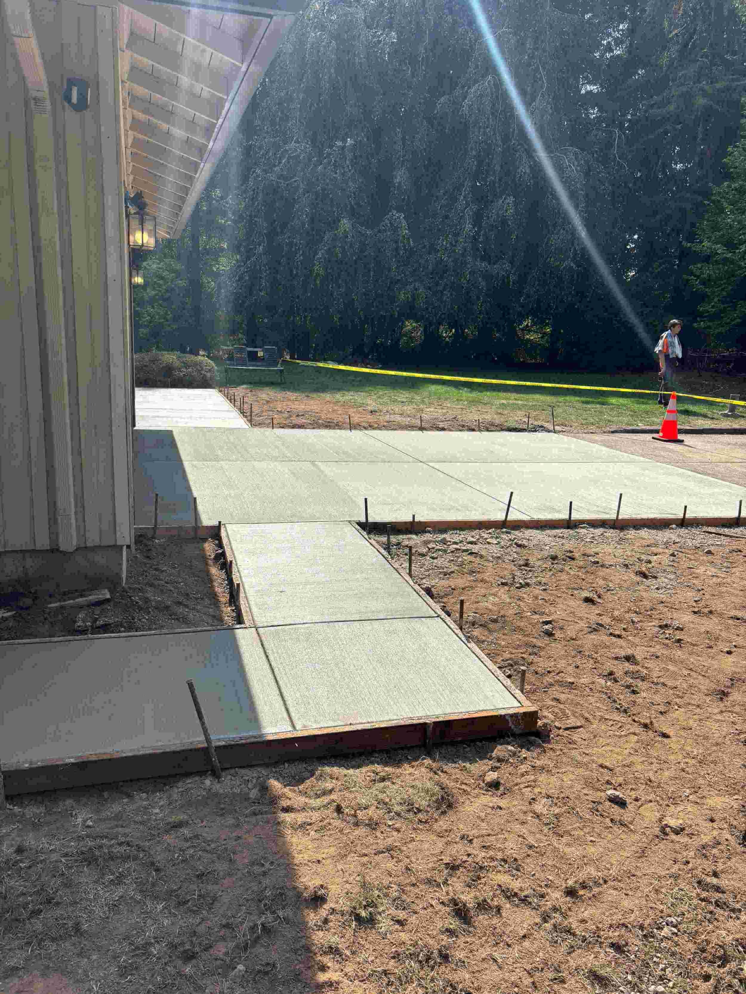 Cement sidewalk being poured in a backyard, with a person walking in the distance, trees and a yellow caution tape in the background.