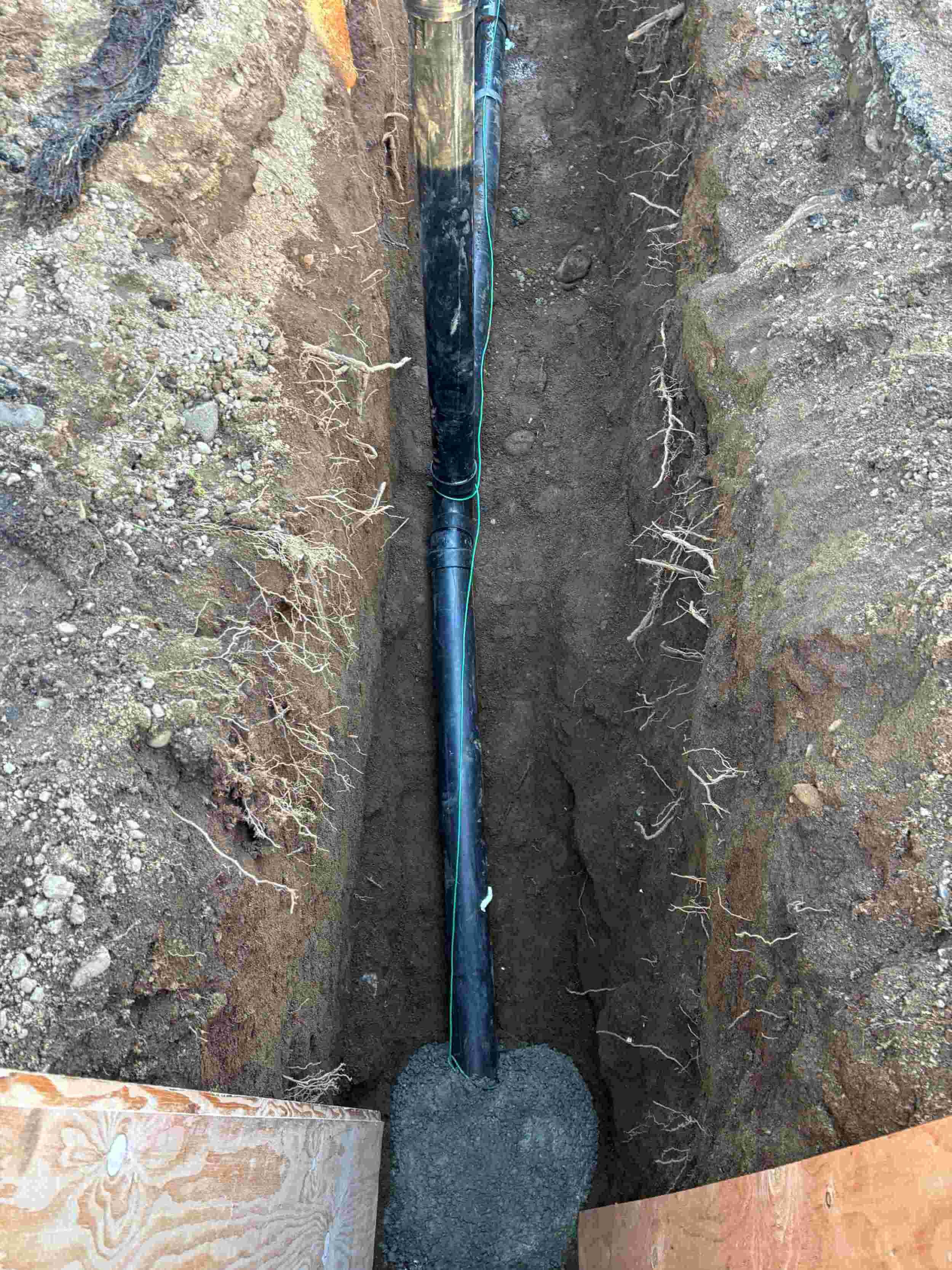 A trench with an underground pipe, some dirt, rocks, and roots, and a black sock of soil at the bottom.