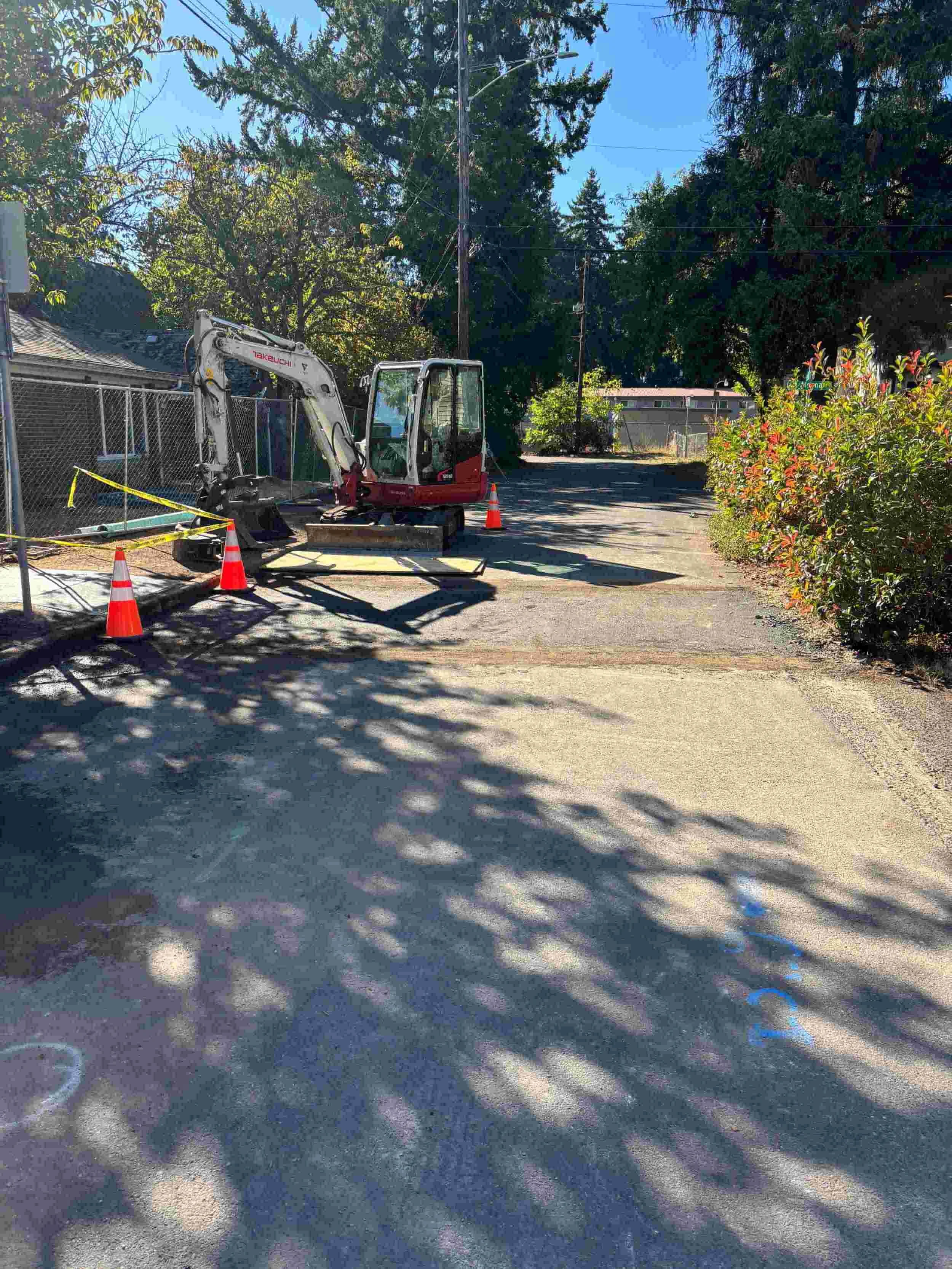 Construction site with a small excavator and orange cones in a shaded alleyway, with trees and bushes on the sides and houses in the background.