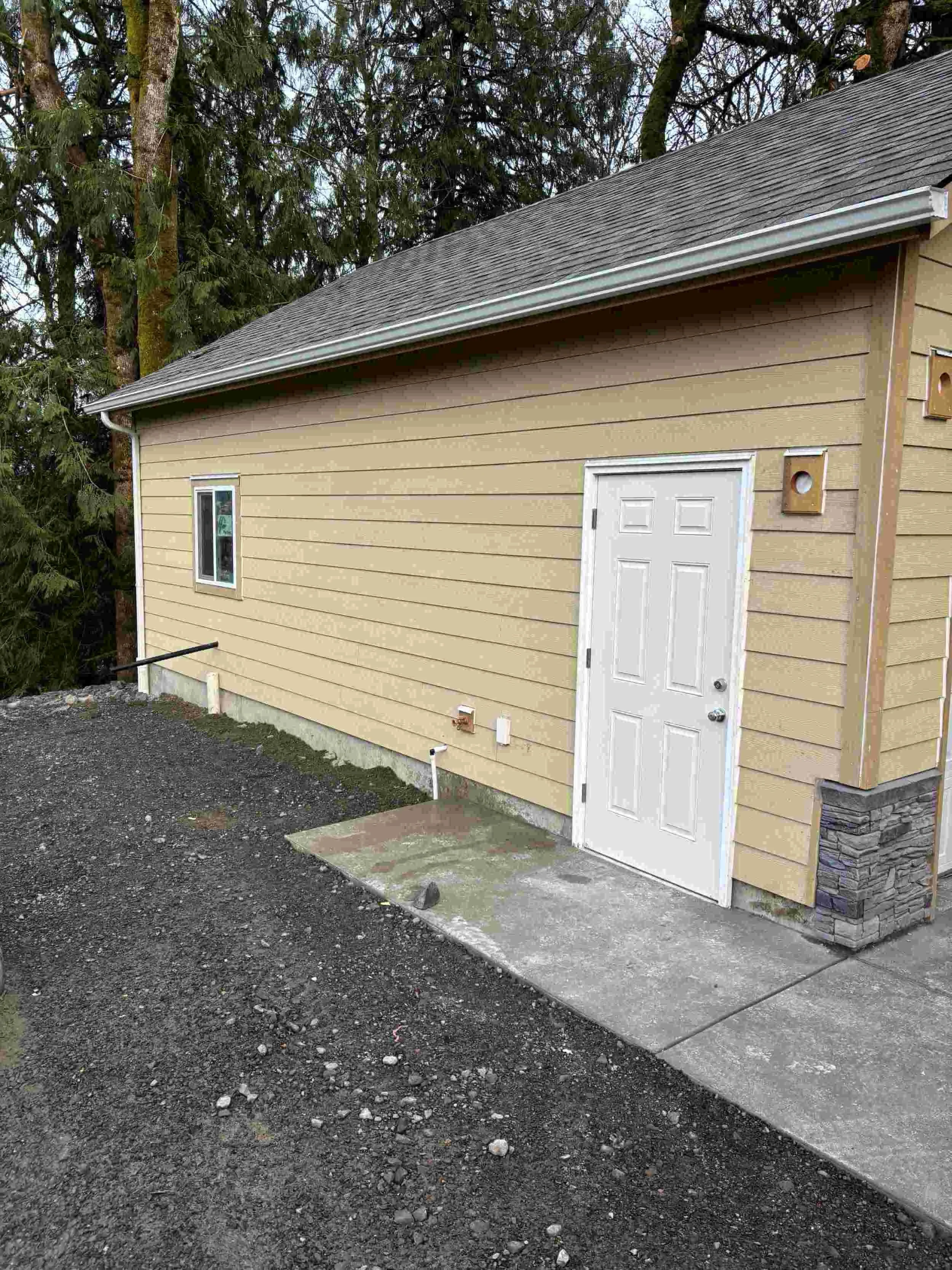 Back of a beige house with a gray roof, a white door, a small window, a vent, and utility pipes, with a gravel and concrete ground and a wooded area in the background.