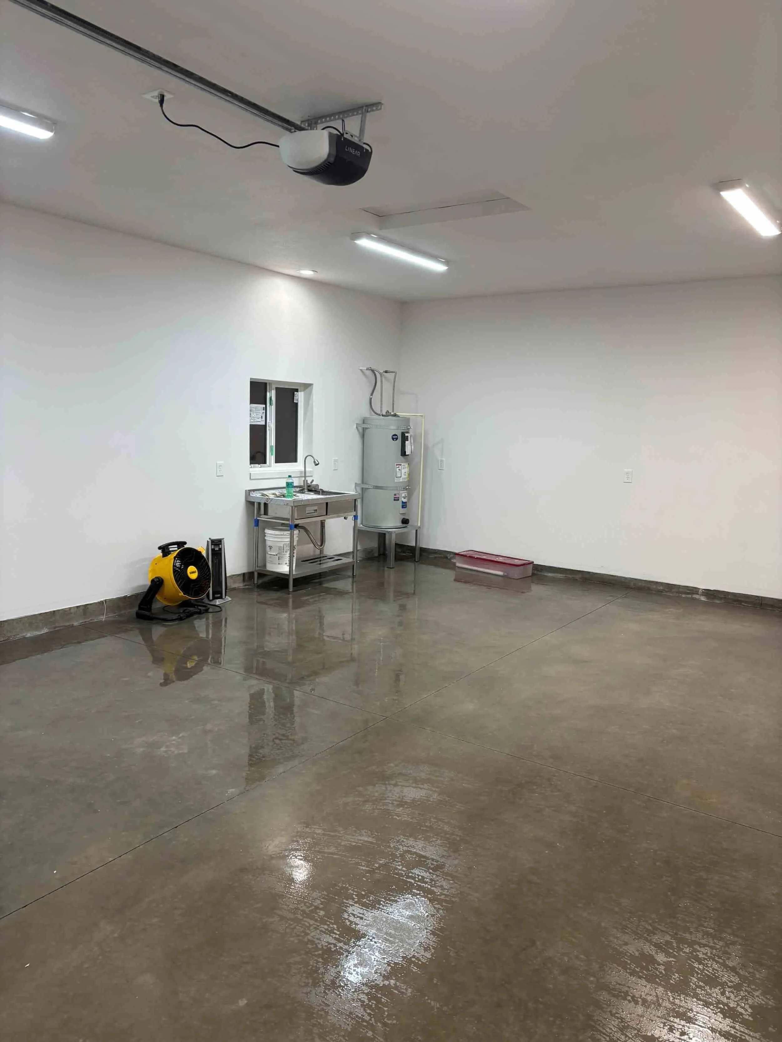 Empty garage or utility room with a wet concrete floor, a water heater, a sink, and cleaning equipment.