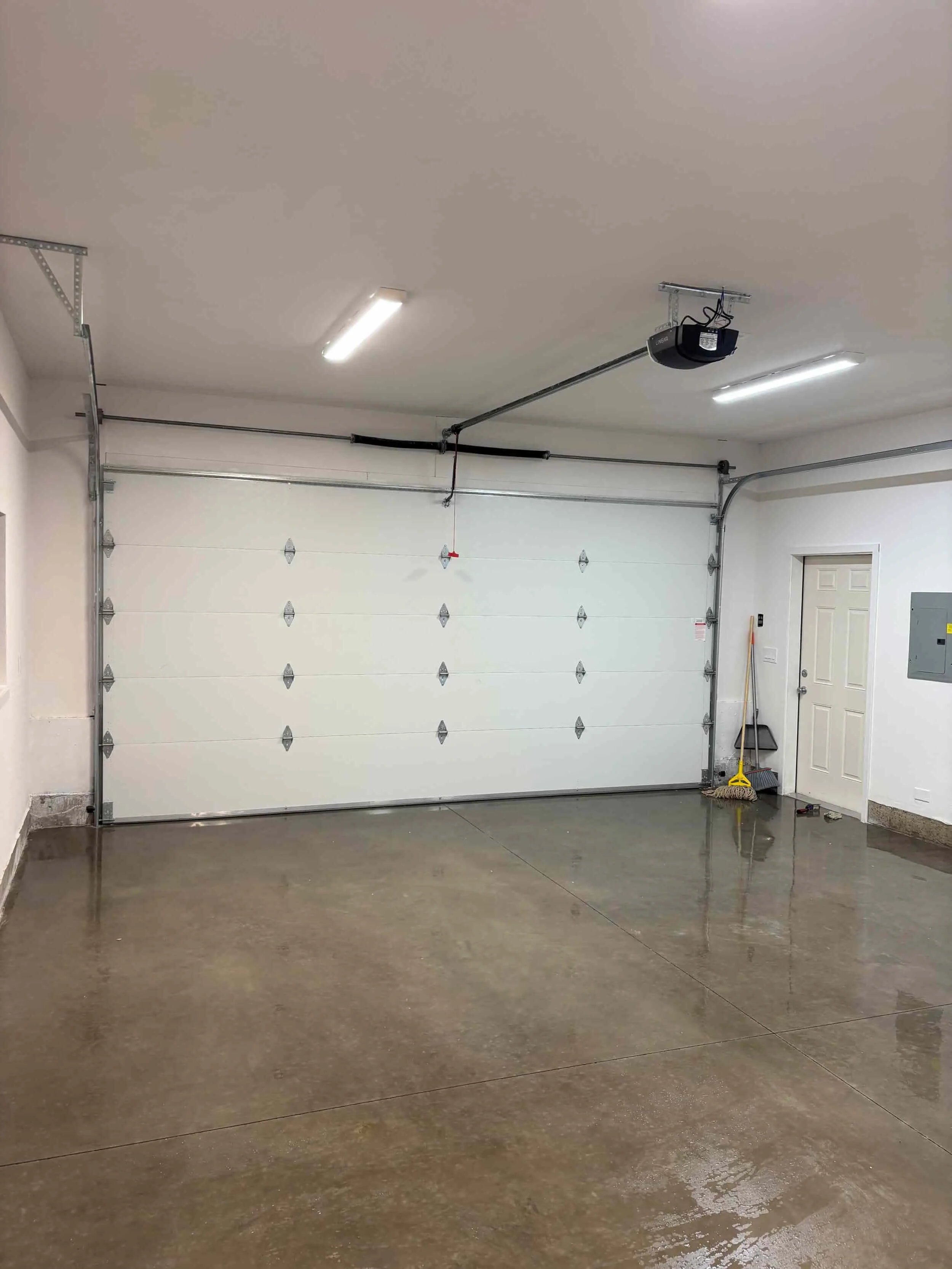 Empty garage with a white overhead door, concrete floor, and cleaning tools near a door on the right side.