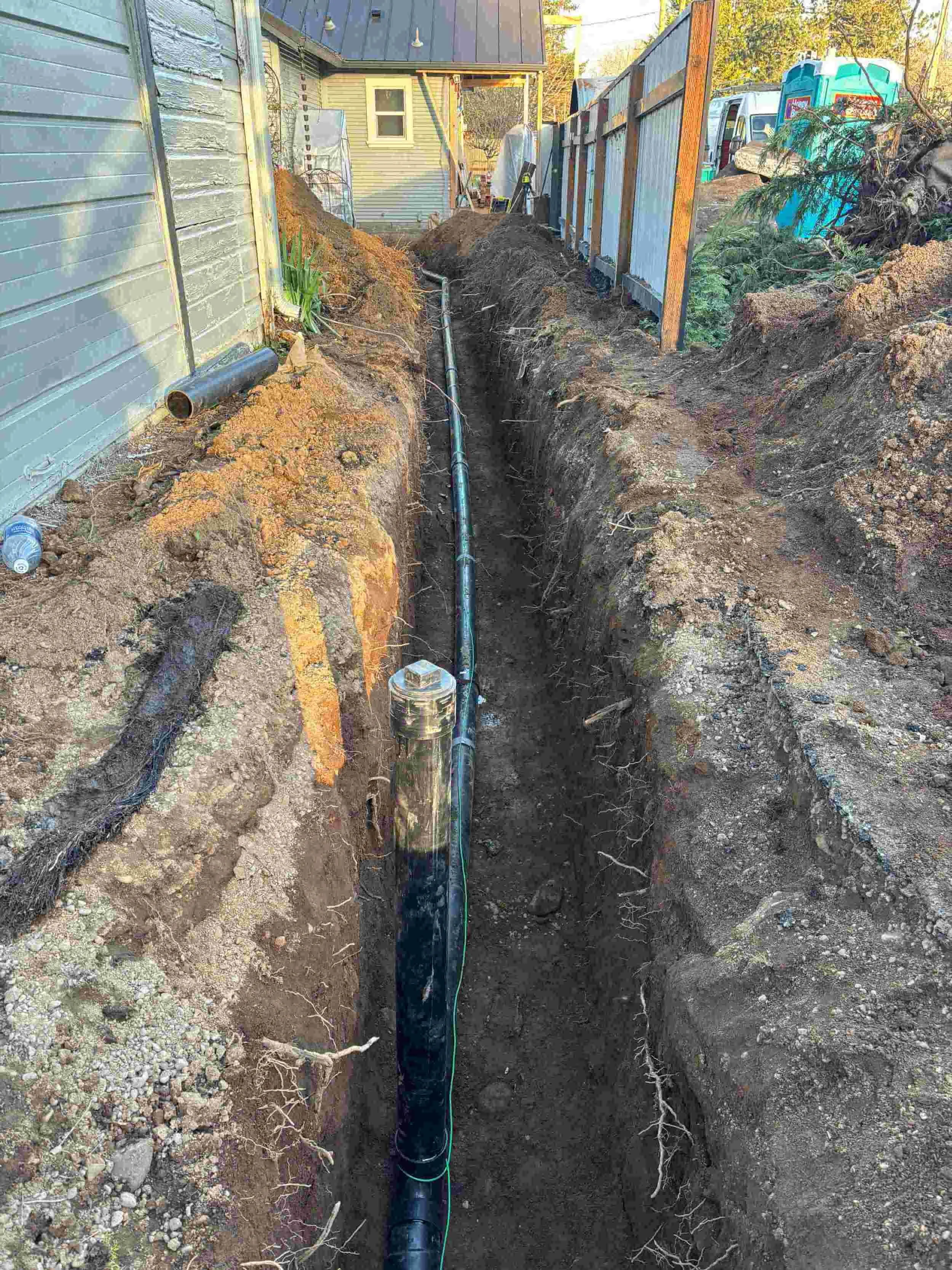 Construction site with a trench showing underground piping and a house in the background.