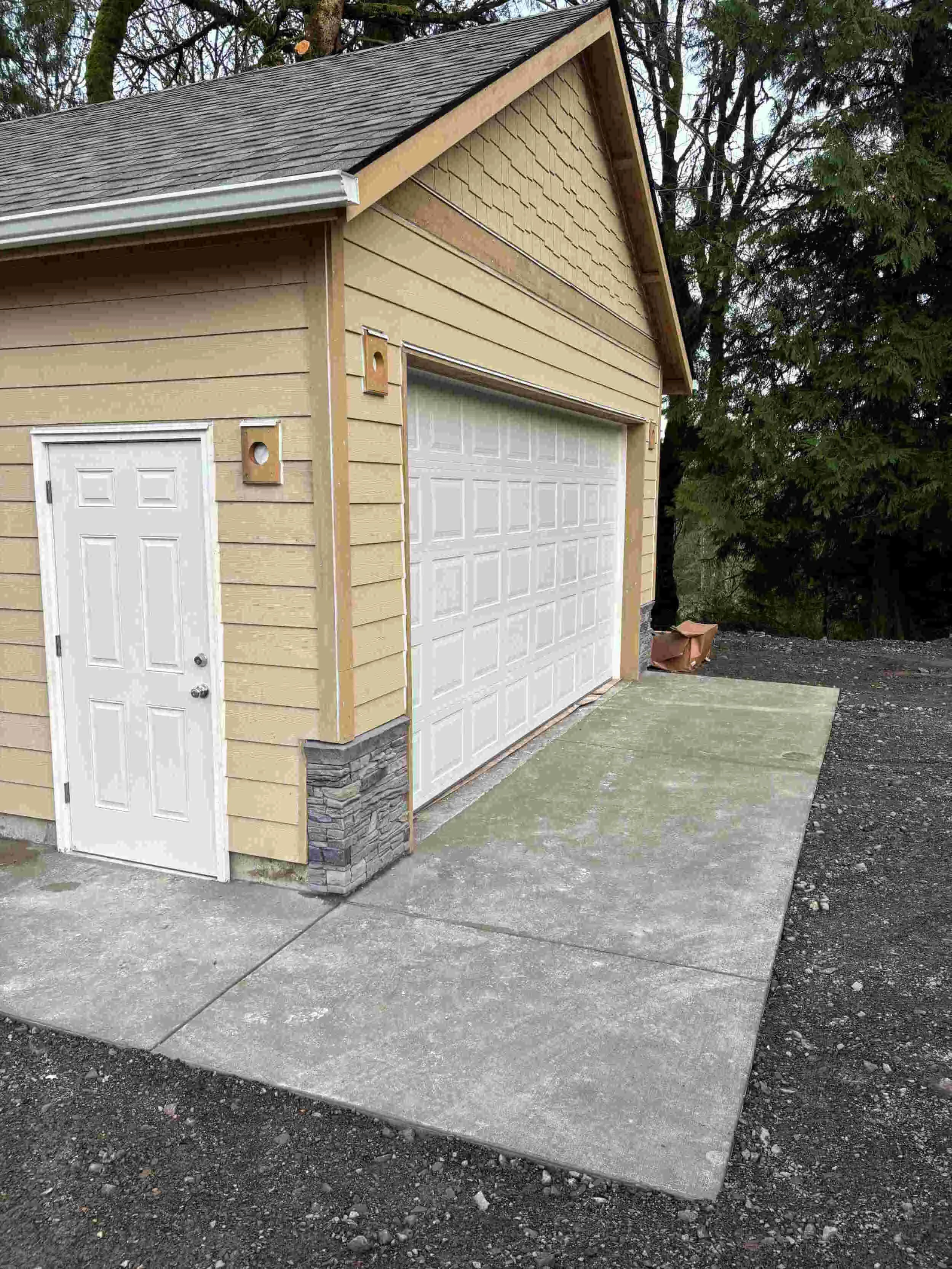 A beige garage with white doors and siding, surrounded by trees, with a concrete driveway and a small white side door.