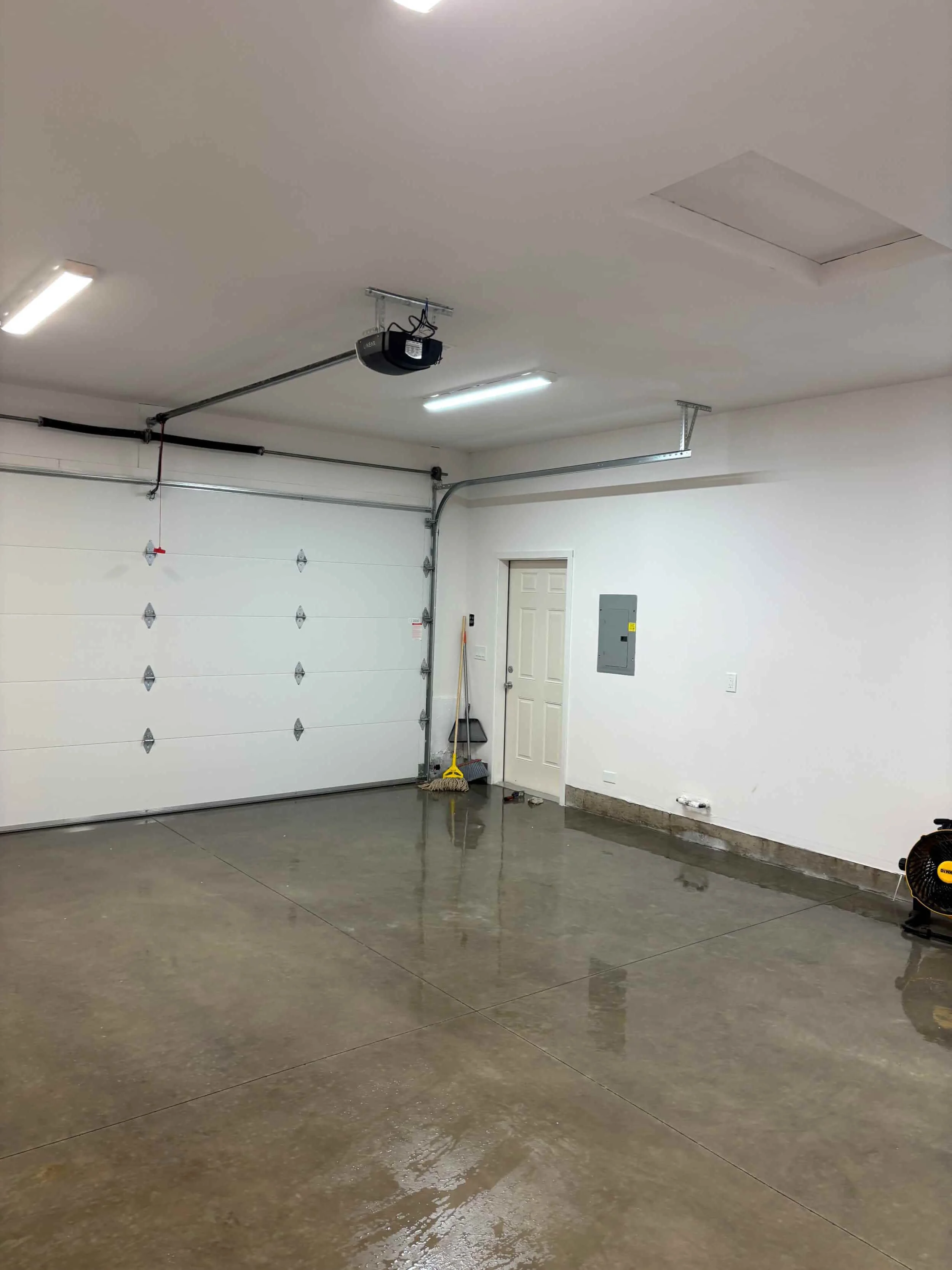 Empty garage with a white roll-up door, concrete floor, and a white wall with a door. Garage tools and equipment are visible, including a broom, dustpan, fan, and overhead garage door opener.