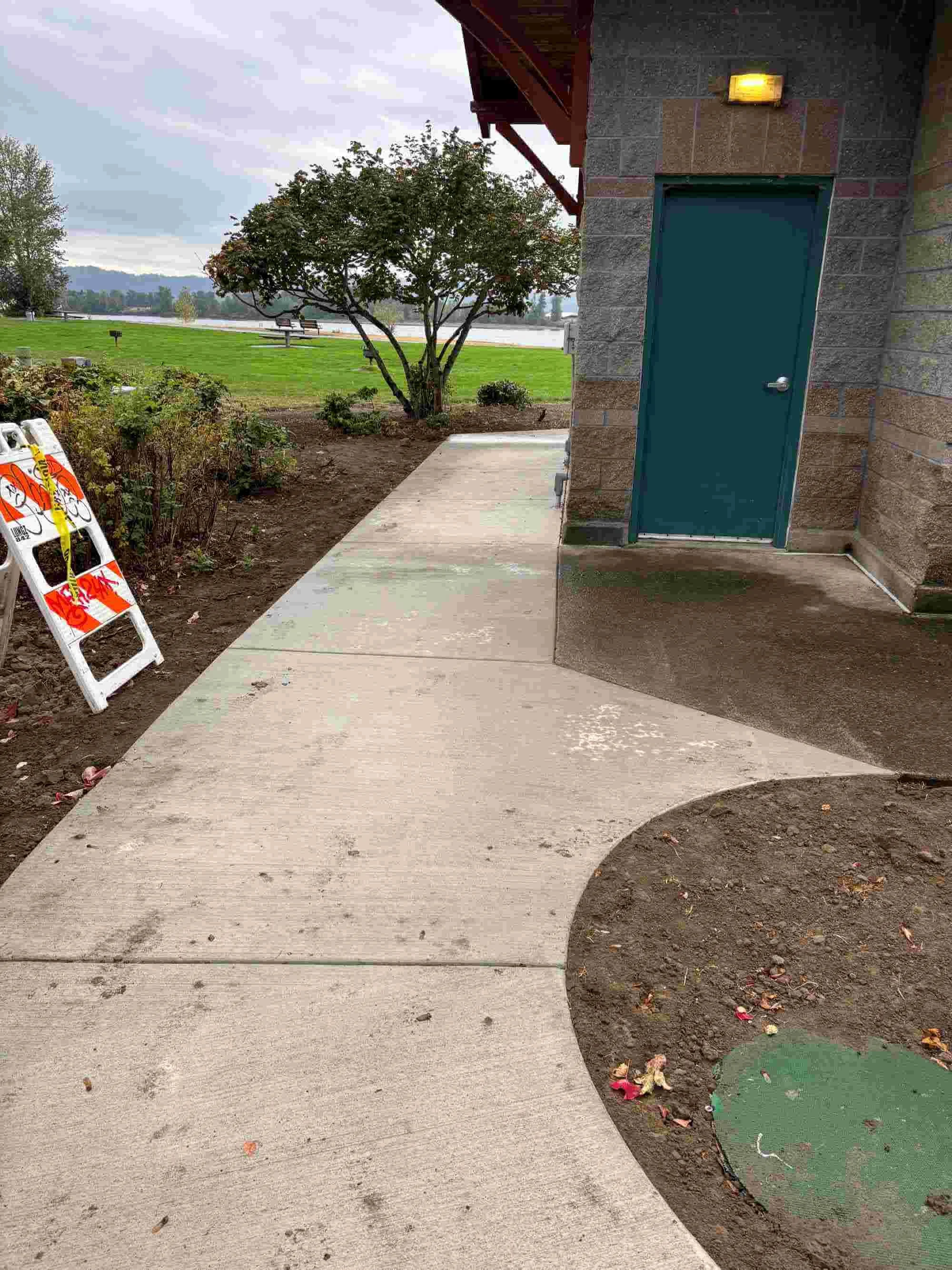 Concrete sidewalk next to a blue door on a brick building, with a yellow light above the door, and a tree in the background near a body of water, overcast sky, and some construction barriers on the side.