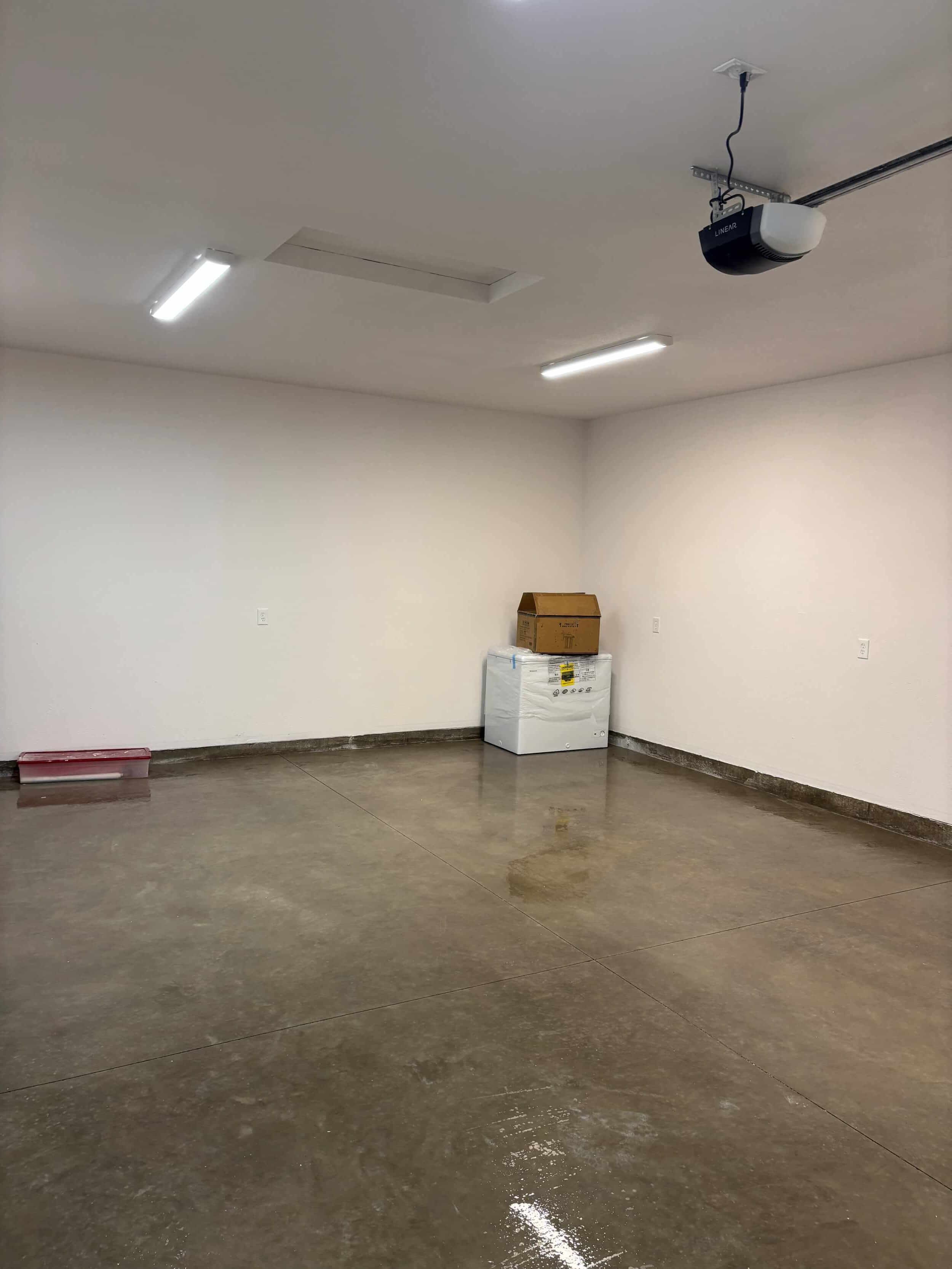Empty garage with white walls, fluorescent lights, and a wet floor; contains a cardboard box and a white appliance wrapped in plastic in the corner.