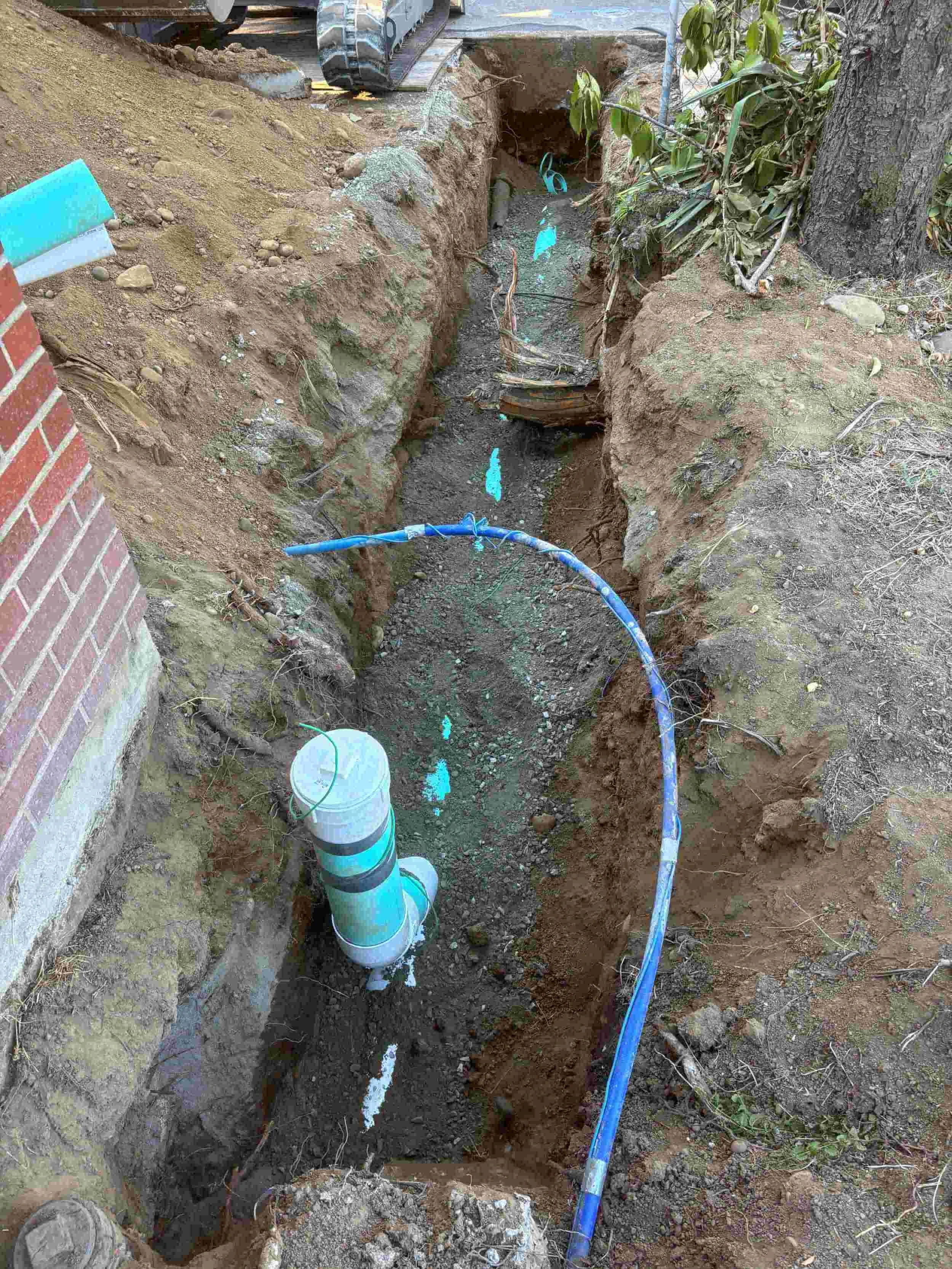Excavation site with a trench containing pipes and green tubing, surrounded by dirt and soil, near a brick wall and a tree, indicating ongoing underground utility work.