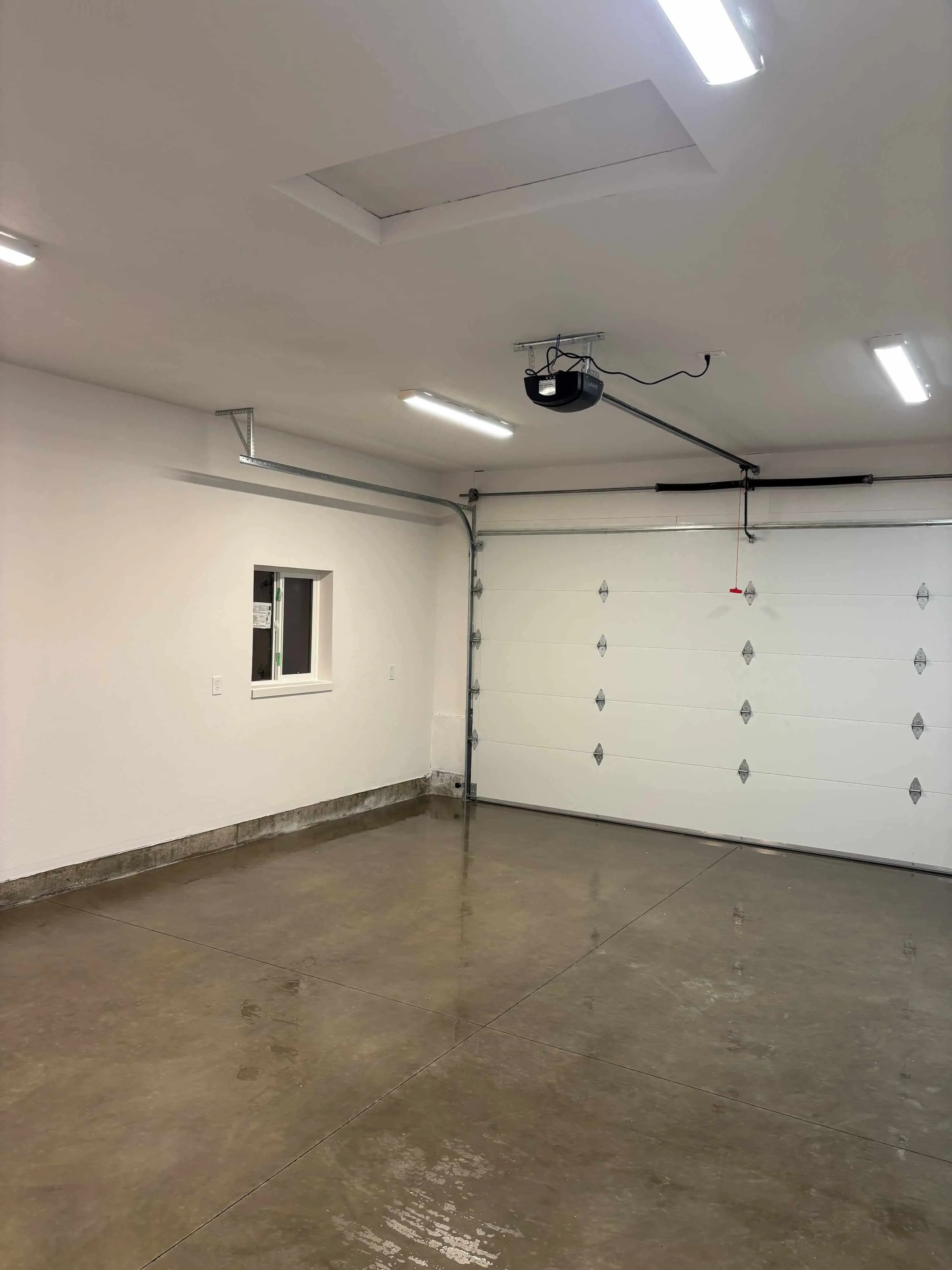 Empty garage with stained concrete floor, white walls, ceiling lights, and a closed white garage door with an automatic opener.